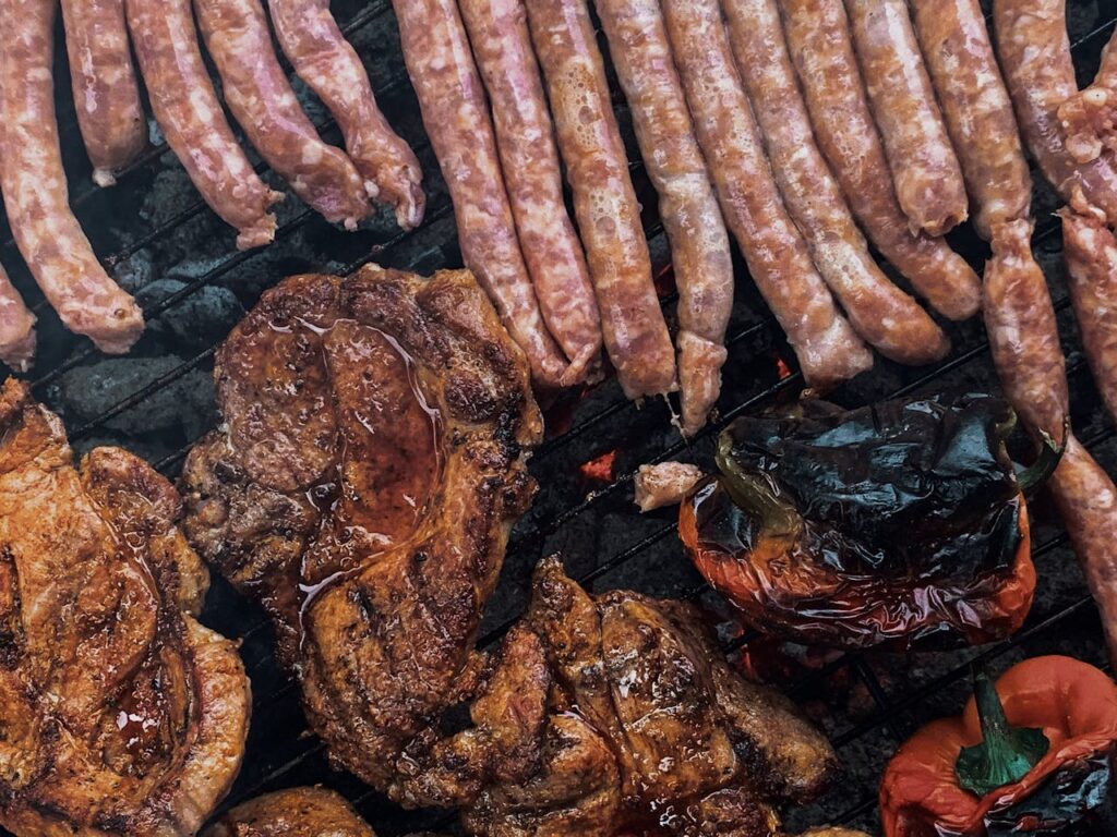 From above yummy steaks and meat sausages roasting on grill grid near organic capsicum during picnic